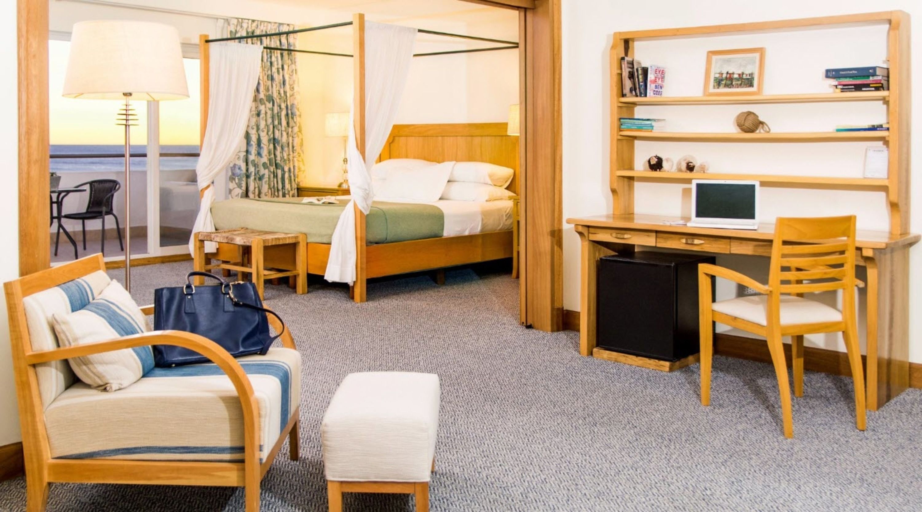 A hotel room with a bed, a desk with a computer, a chair, a small table, and a suitcase.