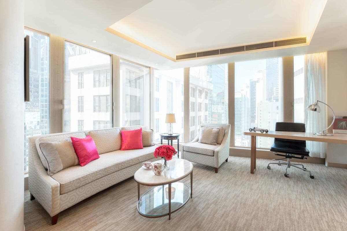 A modern living room with a sofa, a coffee table, a desk, and a chair. Large windows provide a view of the city.