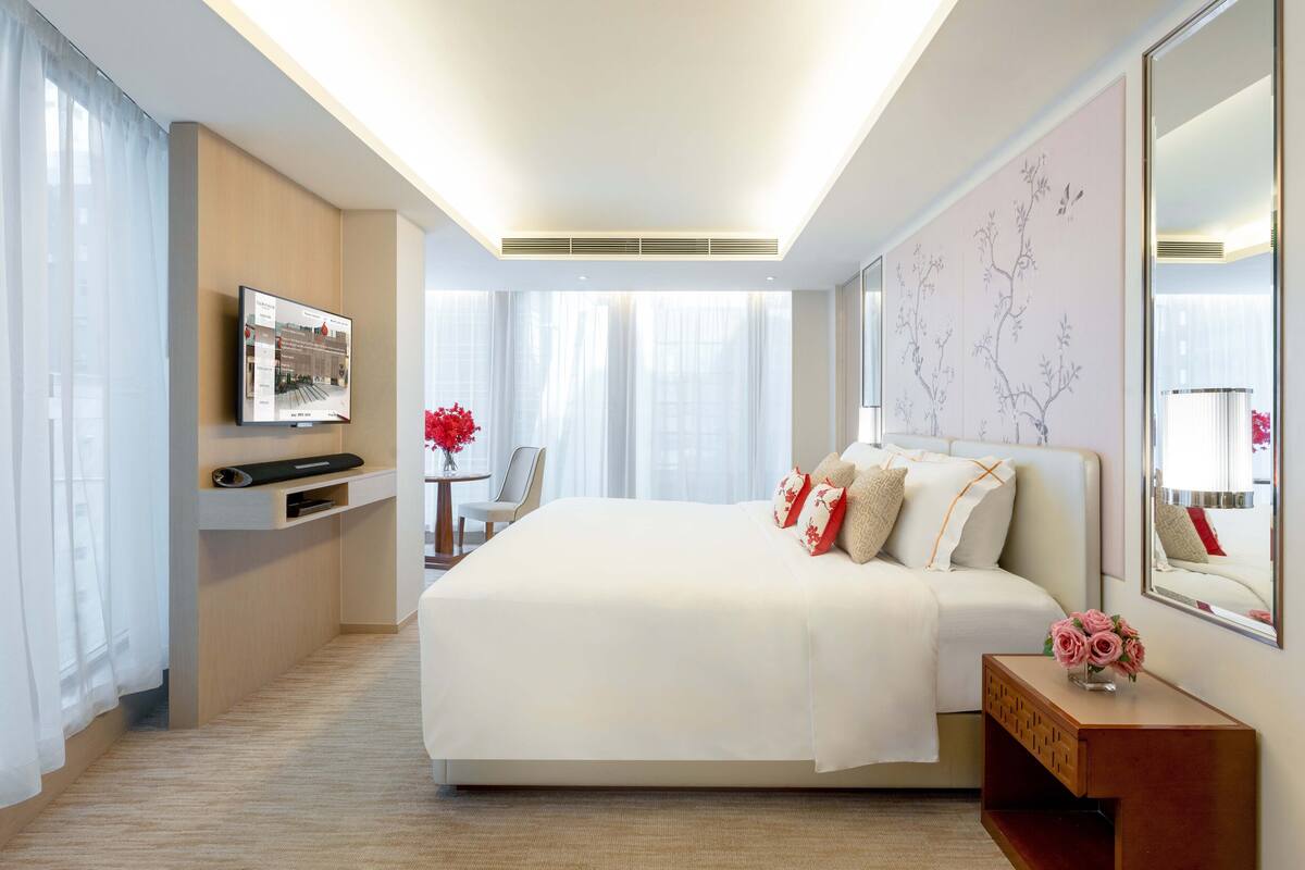 A modern hotel room with a large bed, a flat-screen TV mounted on the wall, a small table with a vase of flowers, and a large window with sheer curtains.