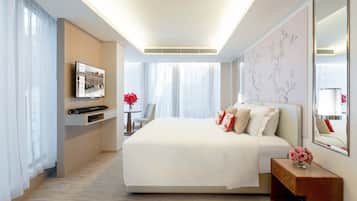 A modern hotel room with a large bed, a flat-screen TV mounted on the wall, a small table with a vase of flowers, and a large window with sheer curtains.