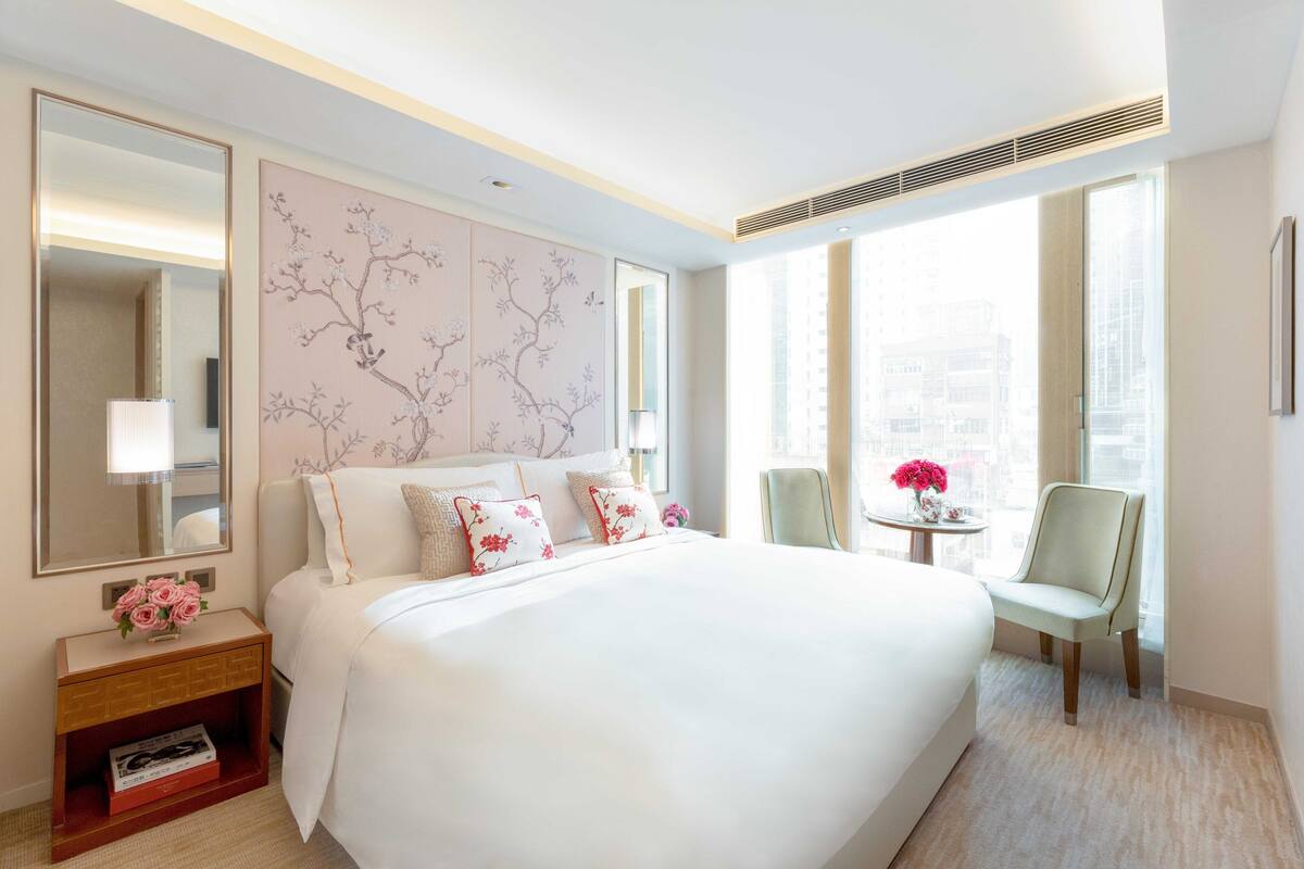 A hotel room with a large bed, a bedside table with a lamp, a chair, and a window with a city view.