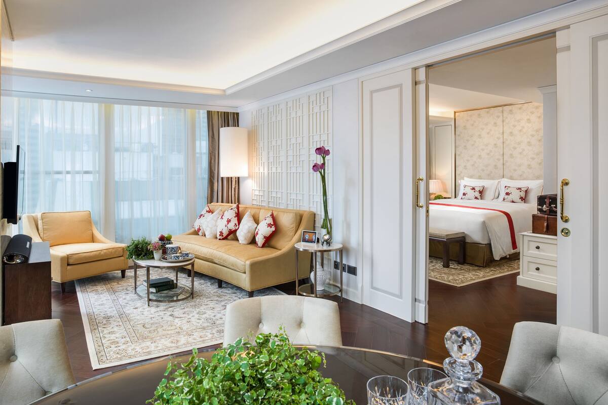 A modern hotel room with a sofa, armchairs, a dining table, and a separate bedroom with a bed and nightstand.