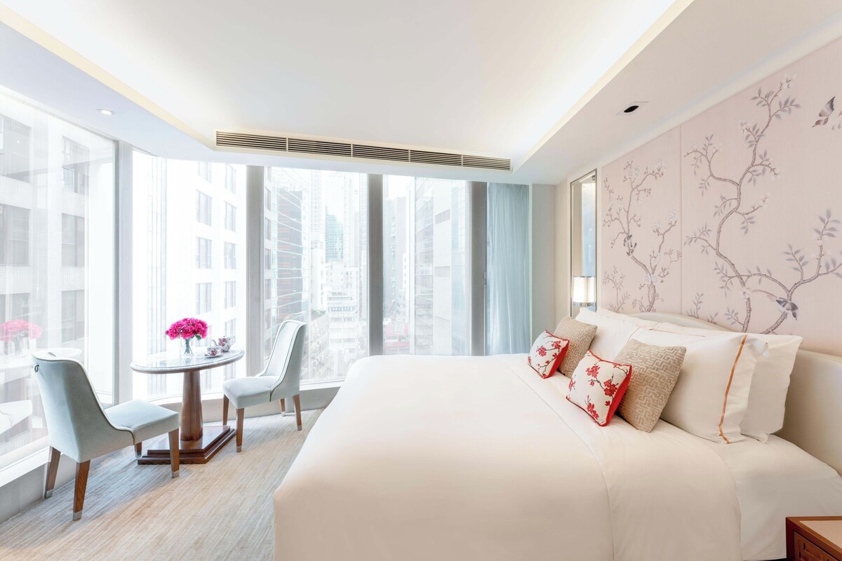 A hotel room with a large bed, a round table with chairs, and a view of tall buildings through large windows.