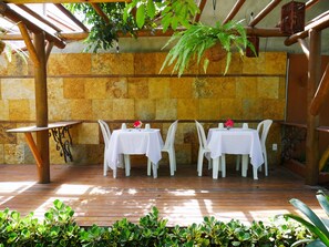 An outdoor patio with wooden flooring, white tables, and chairs, surrounded by stone walls and greenery. - Pousada Enseada dos Golfinhos (Tibau do Sul)
