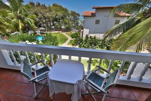 A balcony with a table and two chairs, overlooking a garden with a pool and a house with a red-tiled roof. - Pousada Enseada dos Golfinhos (Tibau do Sul)