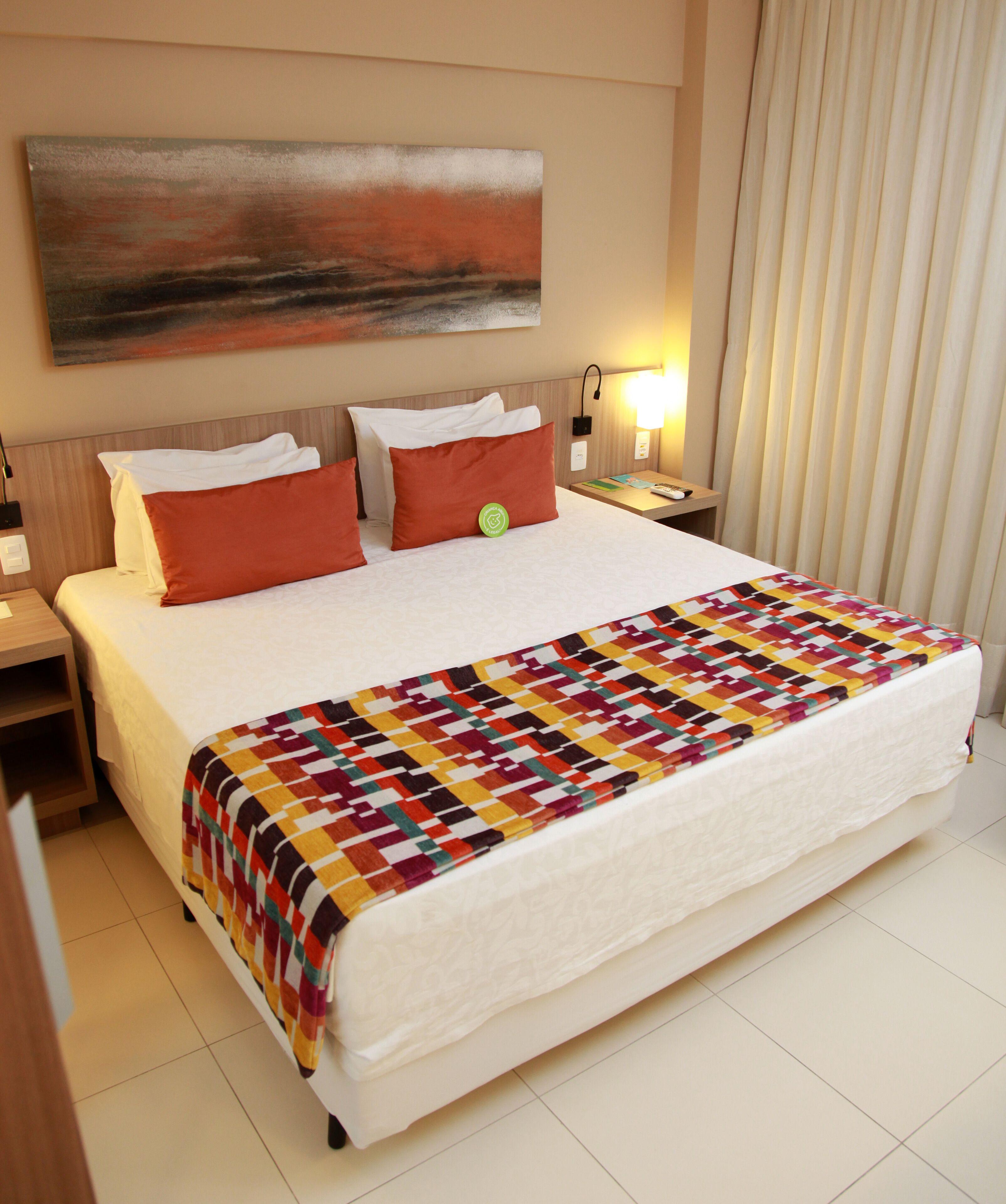 A hotel room with a large bed, a colorful quilt, a painting on the wall, and a nightstand with a lamp.