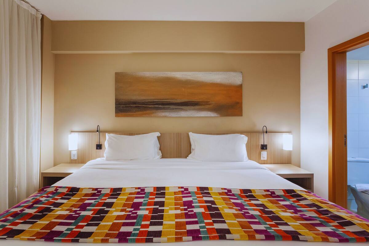 Une chambre d’hôtel avec un grand lit, deux tables de chevet, une couverture colorée, un tableau au mur et une salle de bain visible à travers une porte ouverte.