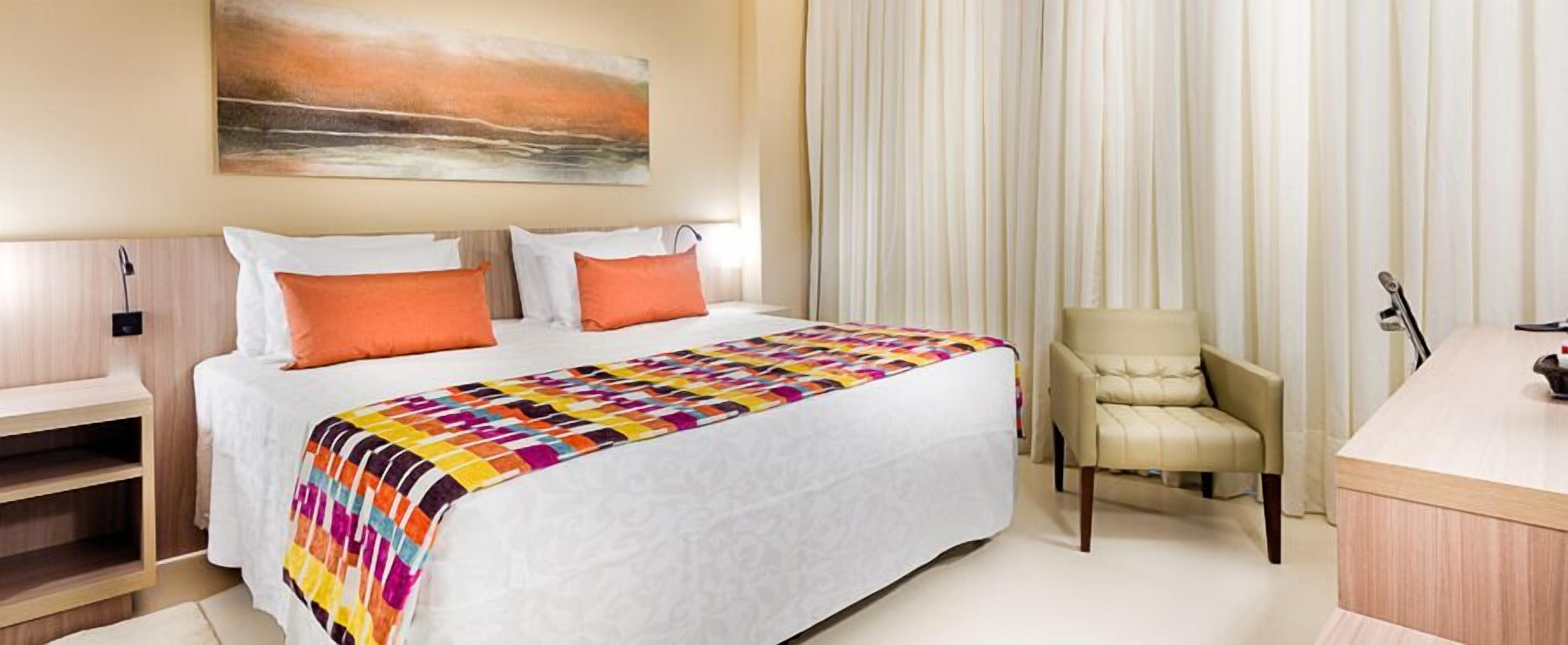 A hotel room with a bed, a chair, a desk, and a painting on the wall.