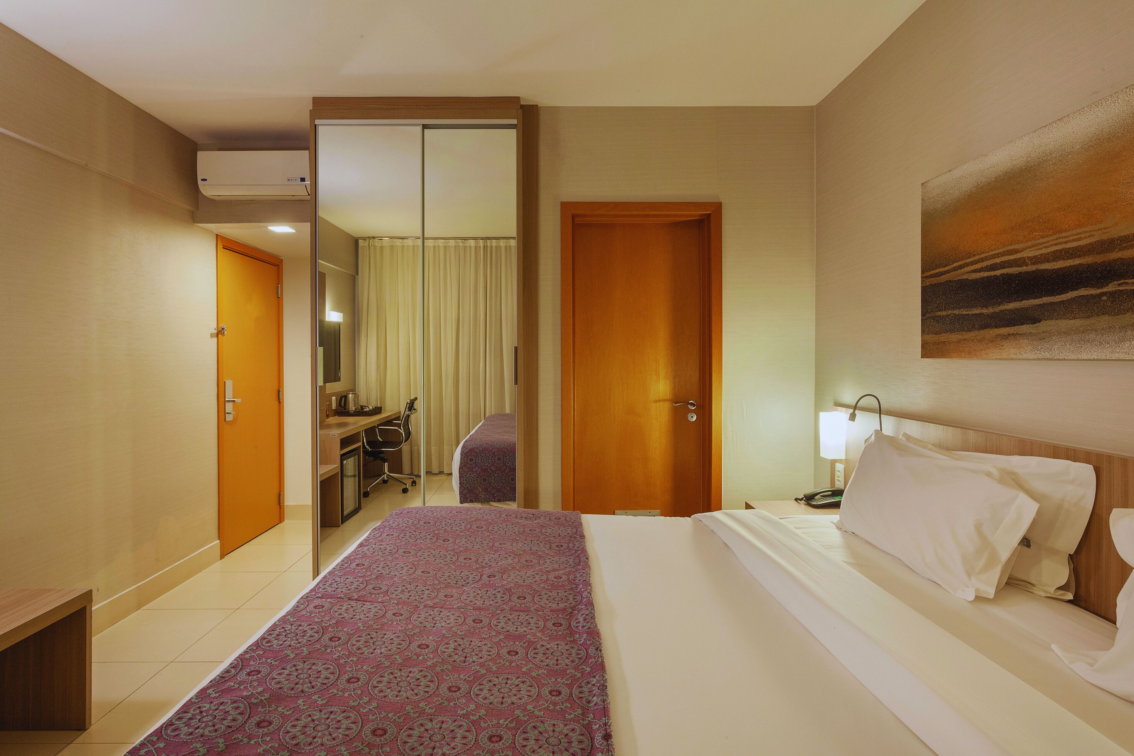 Habitación de hotel con cama, escritorio con silla, espejo y puerta que da a otra habitación.