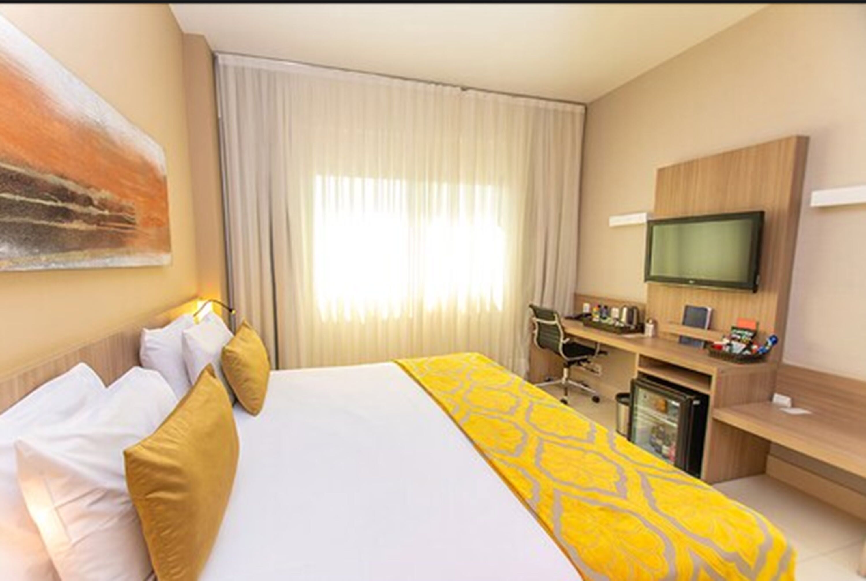 Habitación de hotel con una cama grande, un escritorio con computadora, televisión y ventana con cortinas.