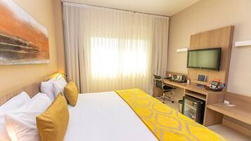 Habitación de hotel con una cama grande, un escritorio con computadora, televisión y ventana con cortinas.