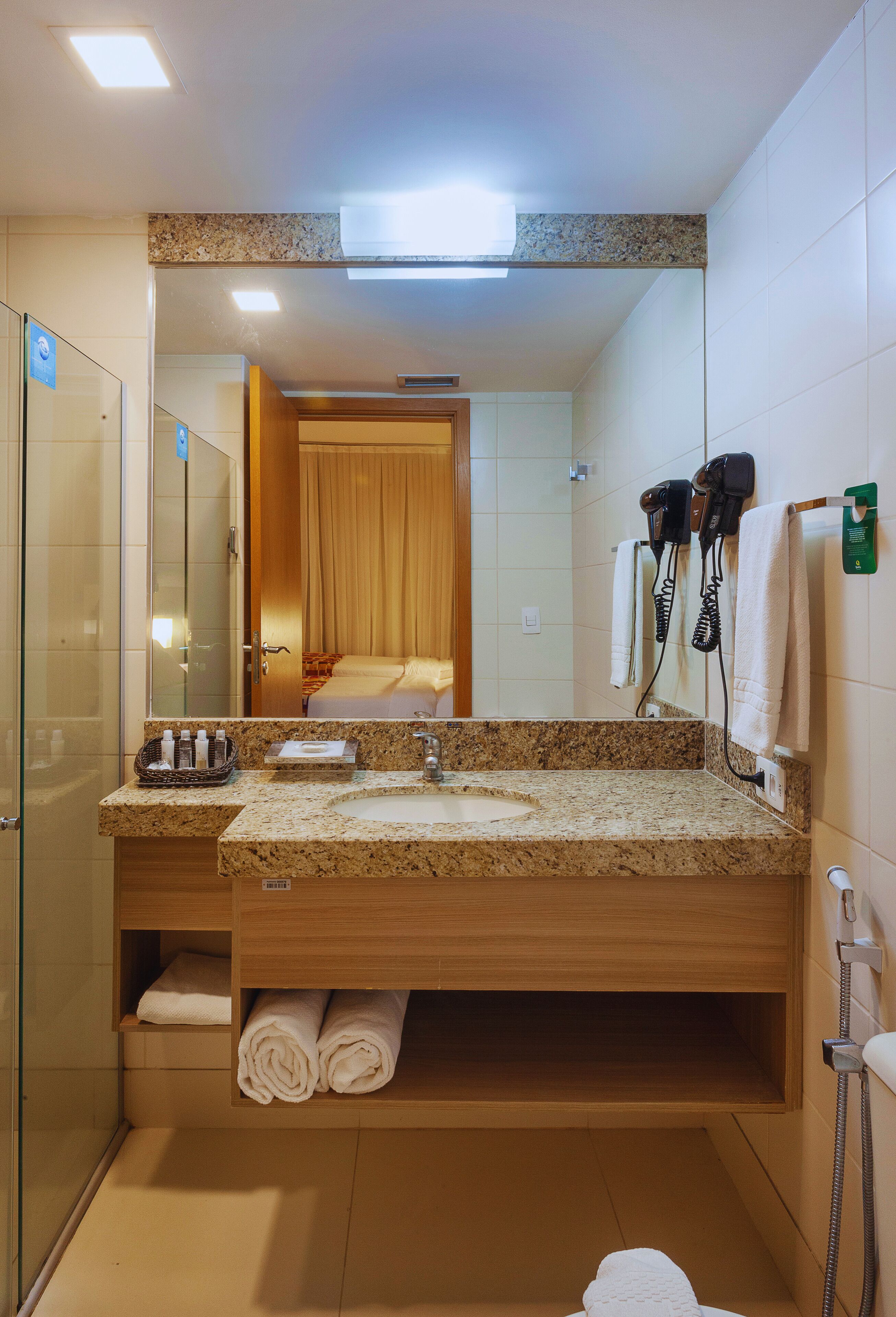 A hotel bathroom with a granite countertop sink, a mirror, a shower area, and a hairdryer on the wall.