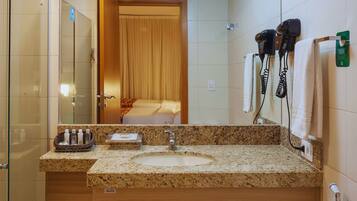 A hotel bathroom with a granite countertop sink, a mirror, a shower area, and a hairdryer on the wall.