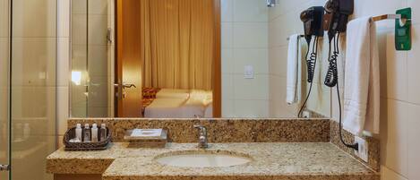A hotel bathroom with a granite countertop sink, a mirror, a shower area, and a hairdryer on the wall.