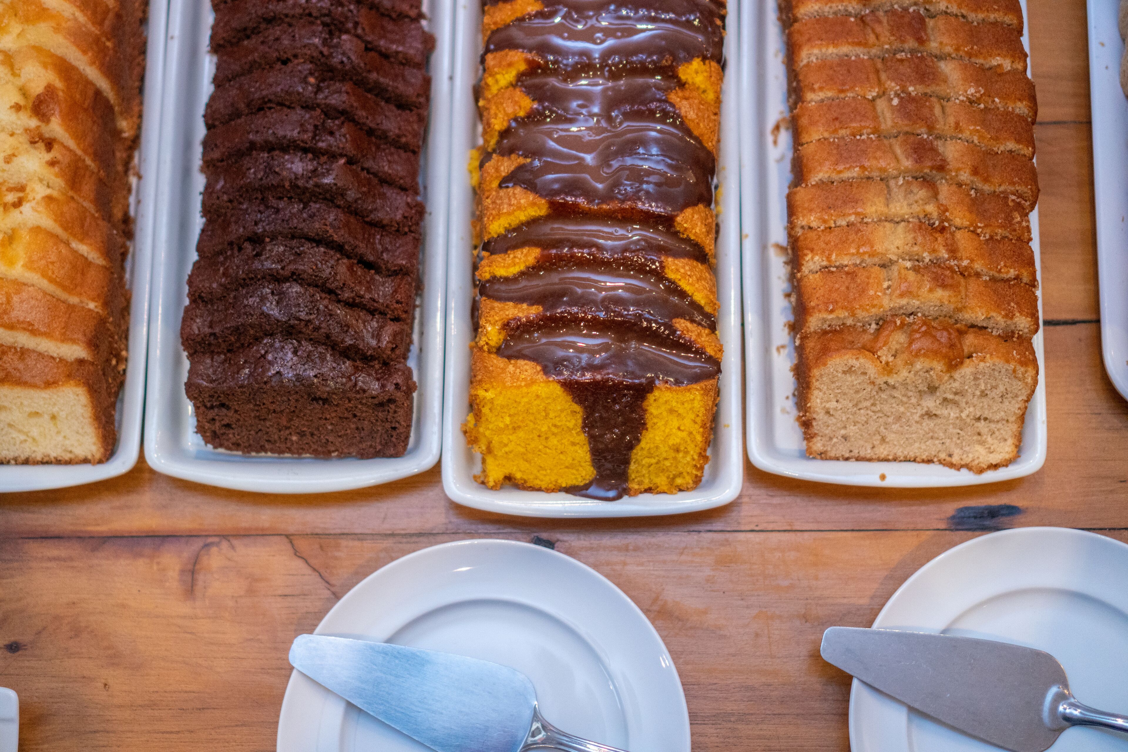 A variety of cakes displayed in white trays on a wooden table.