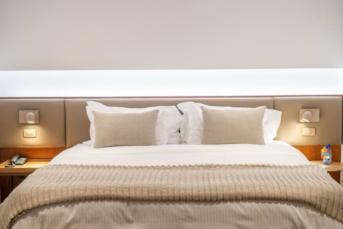 Un lit bien fait, avec une couette et des oreillers de couleur beige, une table de chevet avec une lampe et un téléphone fixé au mur.