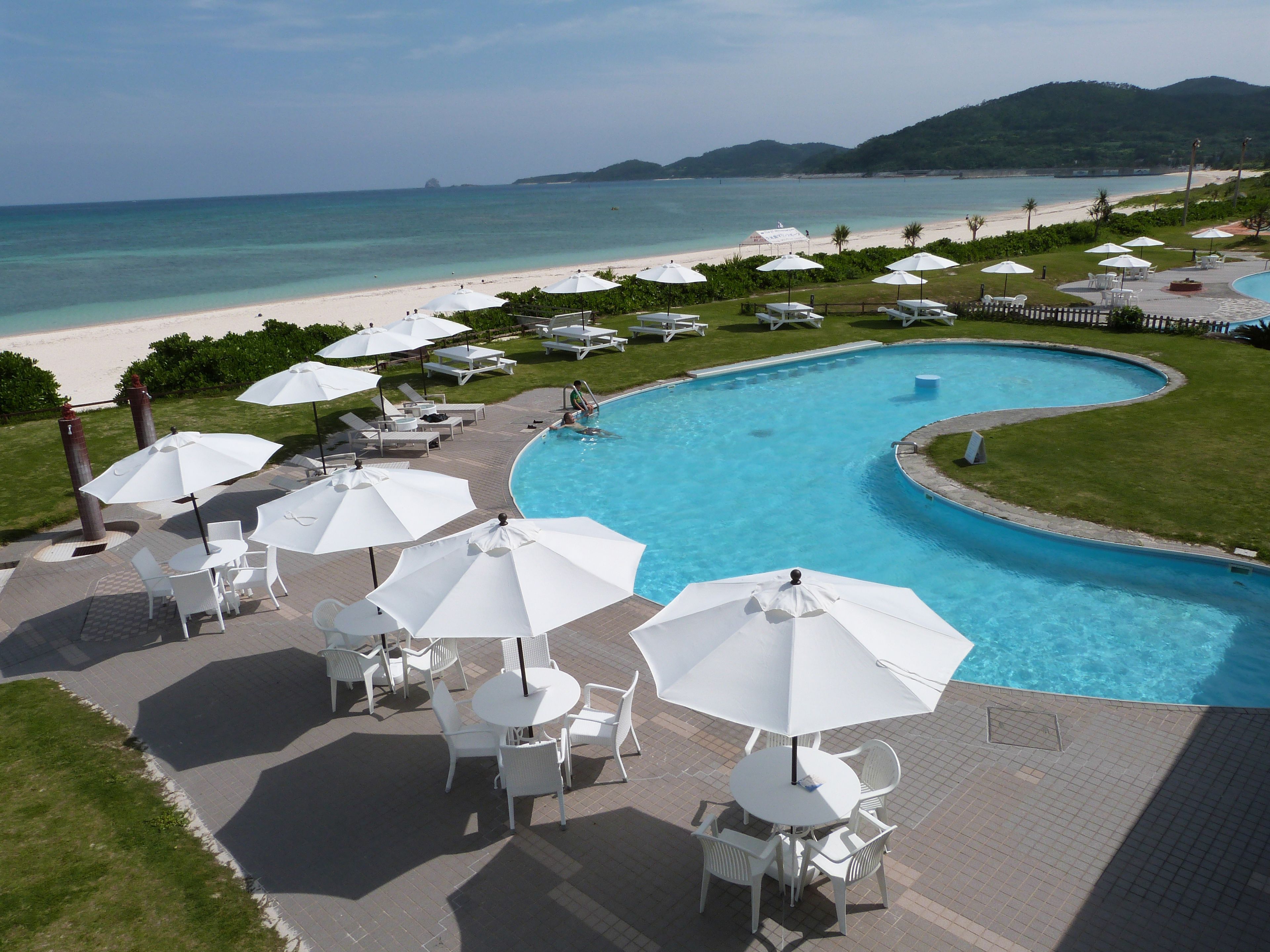 A pool area with white umbrellas, chairs, and a view of the beach and ocean.