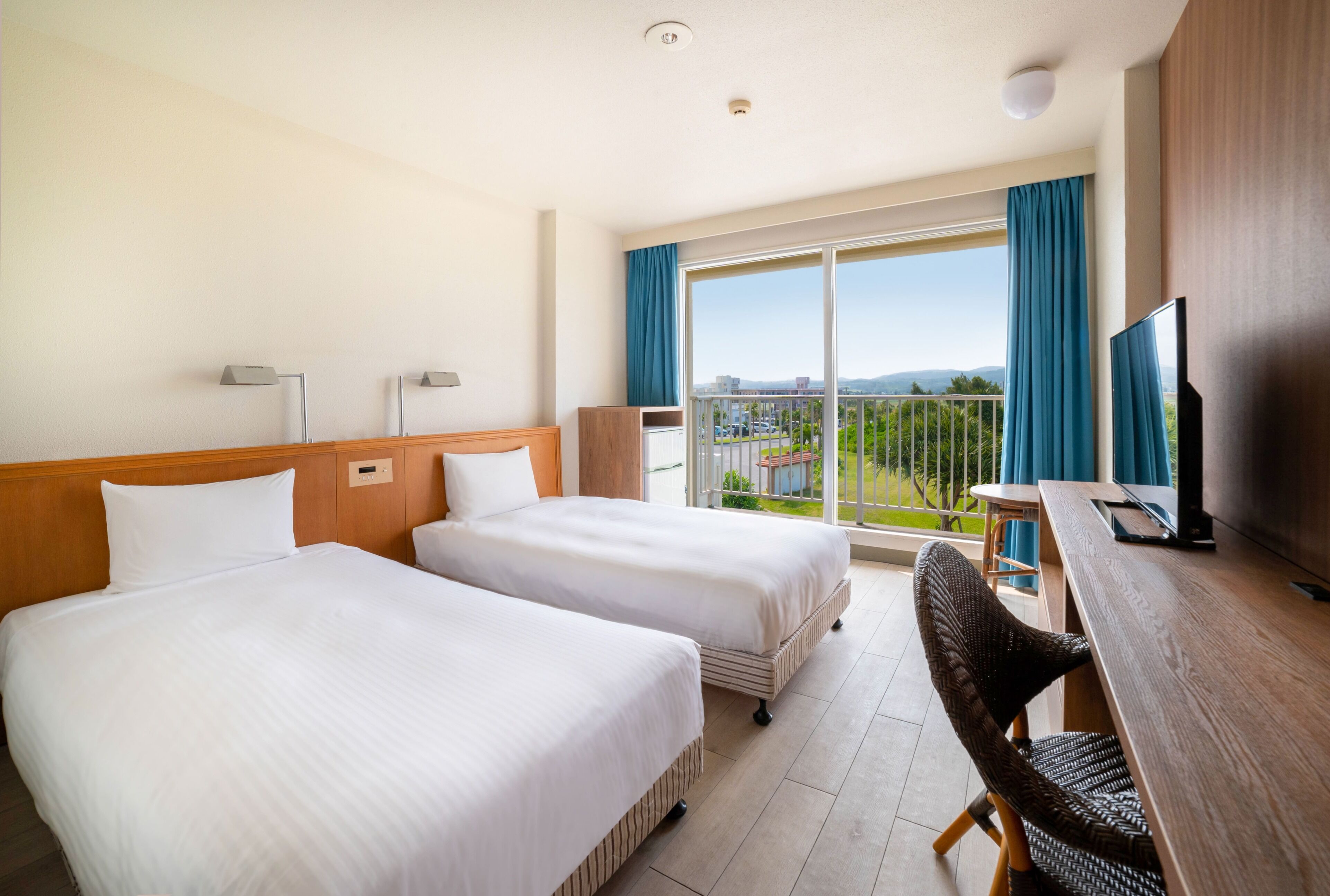 A hotel room with two beds, a desk with a TV, and a balcony with a view.