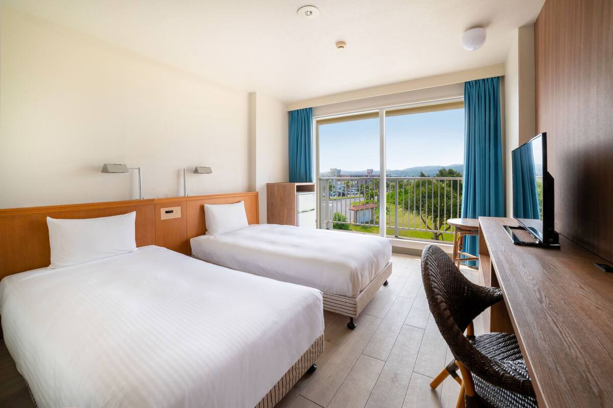 Habitación de hotel con dos camas, un escritorio con televisor y un balcón con vistas.
