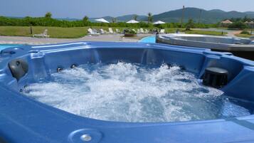 Un jacuzzi con agua burbujeante, rodeado de sillones de descanso y sombrillas, con una piscina y montañas al fondo.