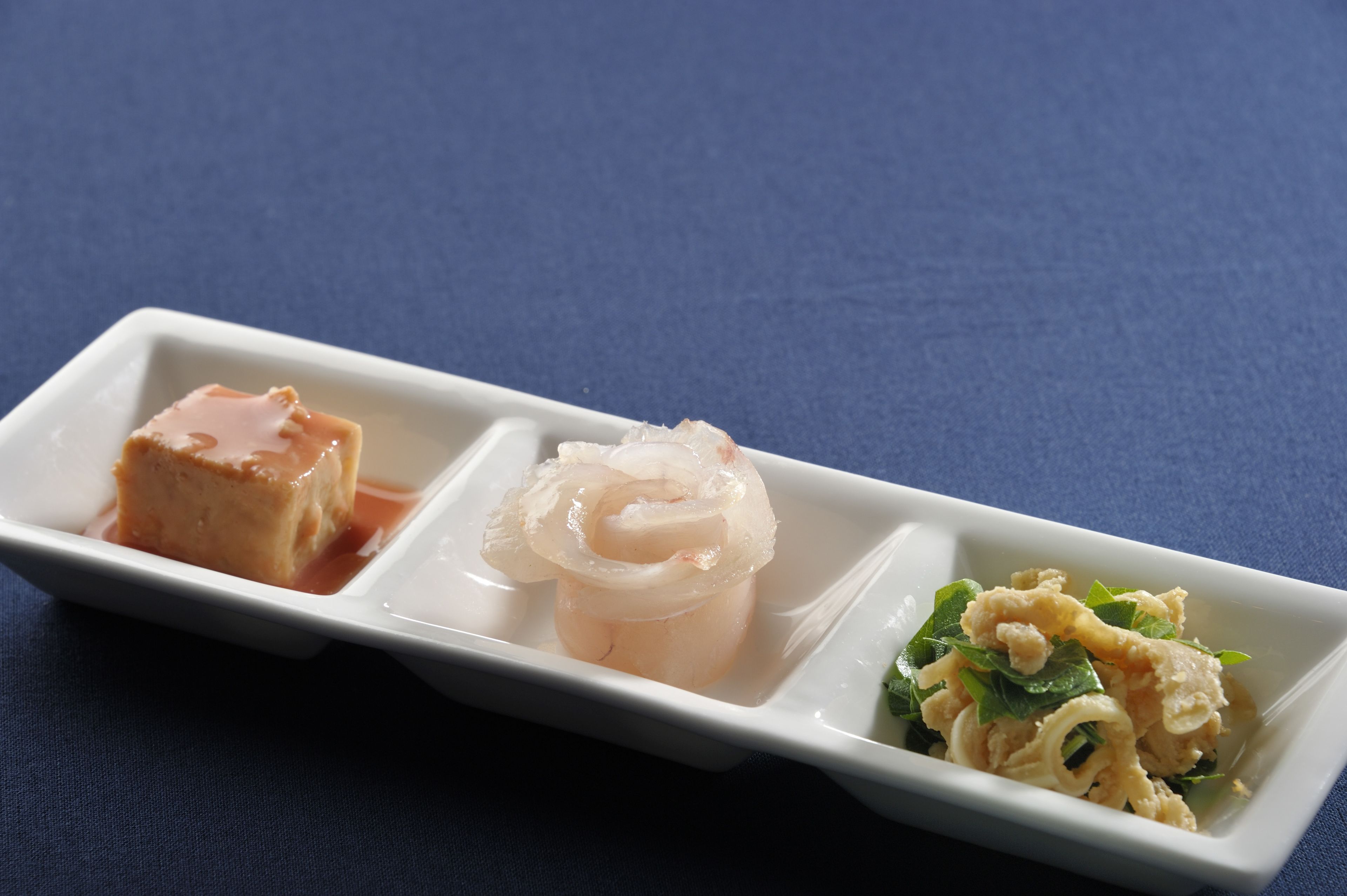 Une assiette rectangulaire blanche sur laquelle sont disposés trois types d’aliments différents : un cube de tofu, un morceau de sashimi et un plat contenant des nouilles et des légumes verts.