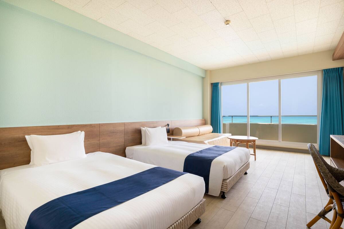Habitación de hotel con dos camas, una mesita, una silla y un ventanal con vista al mar.