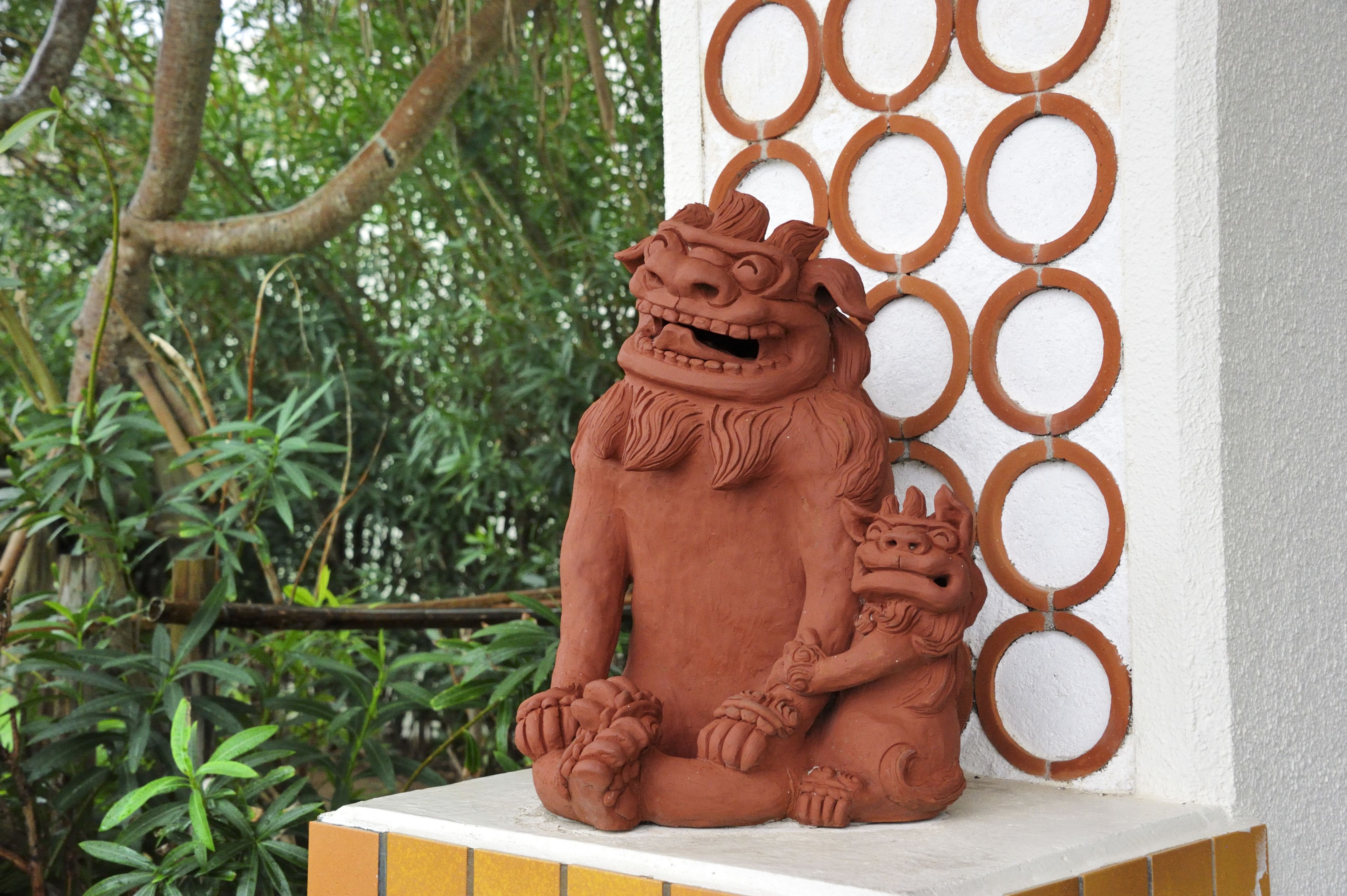 Una estatua de león de terracota con la figura de un niño, frente a una pared blanca con diseños circulares, rodeada de follaje.