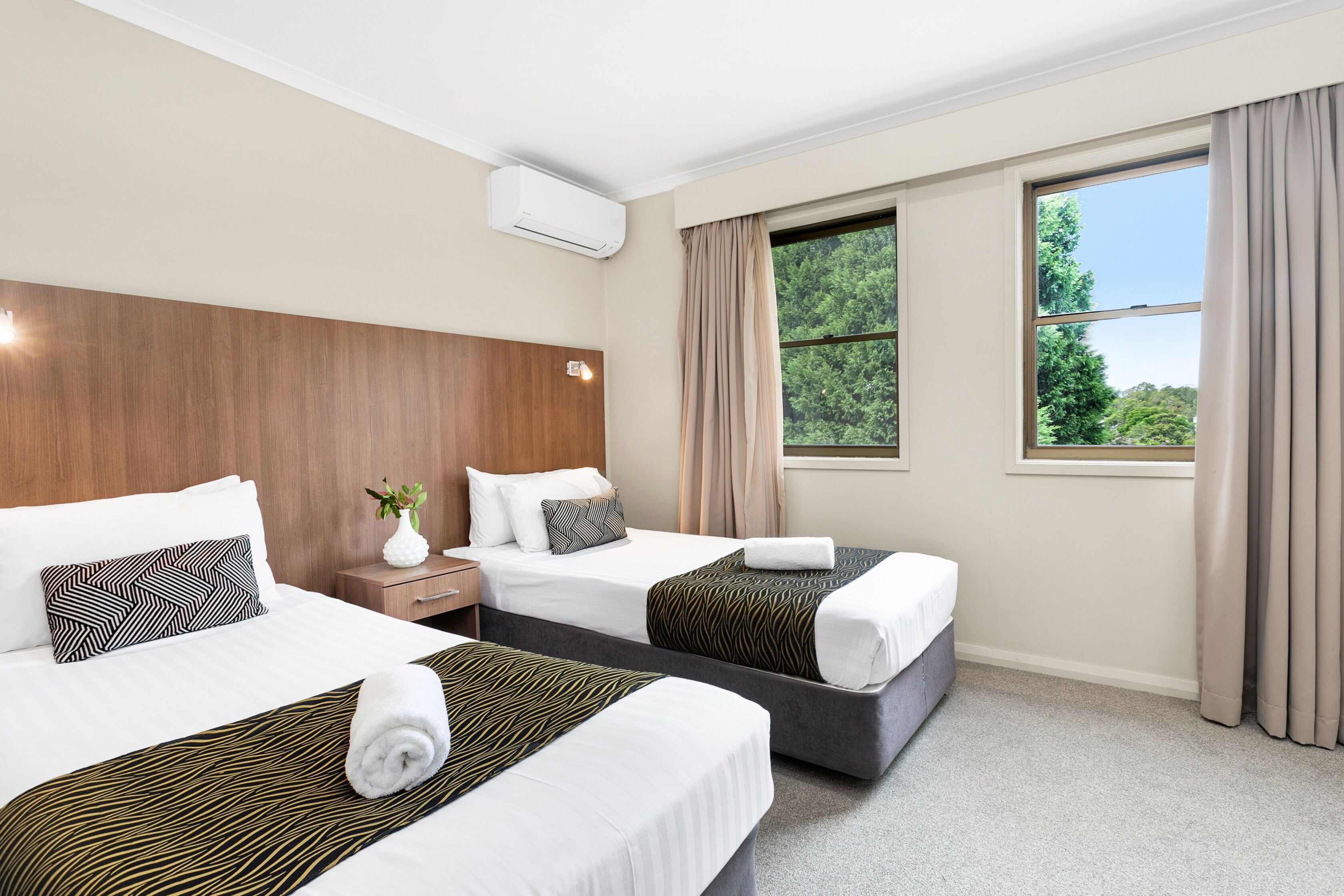 A hotel room with two beds, a wooden headboard, a window with curtains, and an air conditioning unit.