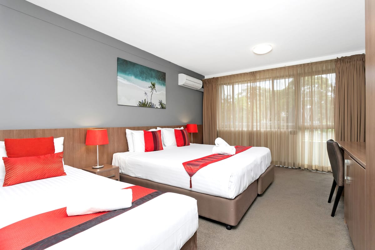Een hotelkamer met twee bedden, een bureau, een stoel, een raam met gordijnen, een airconditioning en een schilderij aan de muur.