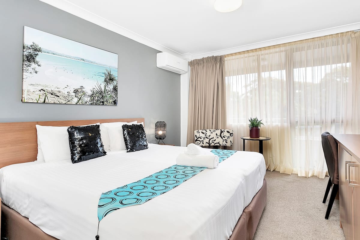 Hotelkamer met een groot bed, een bureau, een stoel, een bijzettafeltje met een plant en een schilderij van een strand.