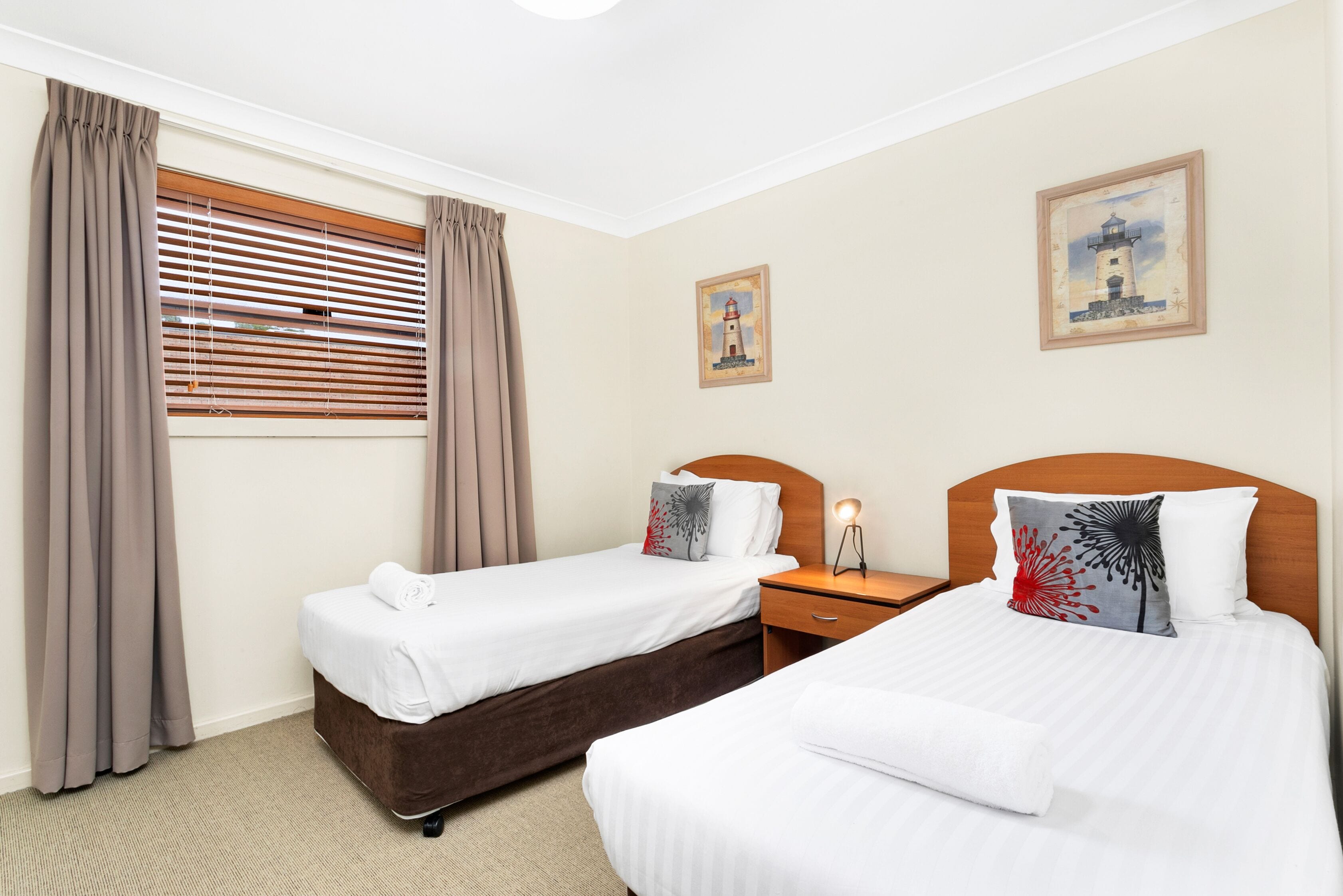 A hotel room with two single beds, a nightstand, a lamp, a window with blinds, and two wall art pieces.