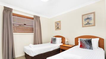 A hotel room with two single beds, a nightstand, a lamp, a window with blinds, and two wall art pieces.