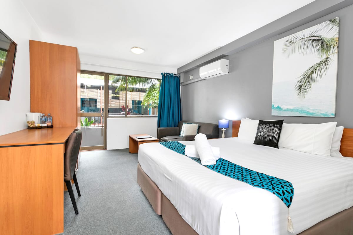 Hotelkamer met een bed, bureau en stoel. Er is een raam met uitzicht op palmbomen en een gebouw.
