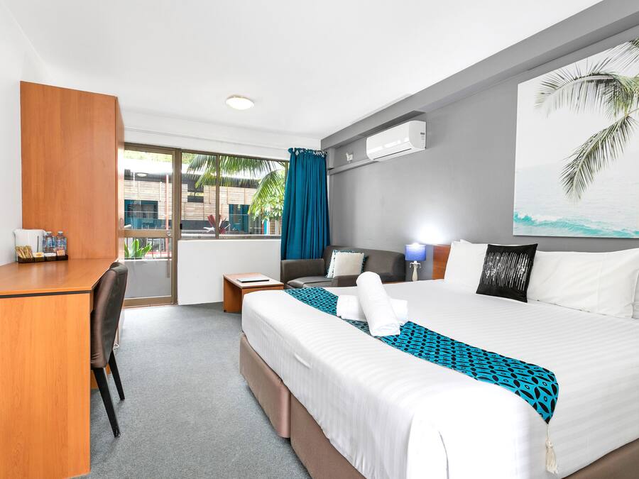 Habitación de hotel con cama, escritorio y silla. Se ve una ventana con vista a palmeras y un edificio.