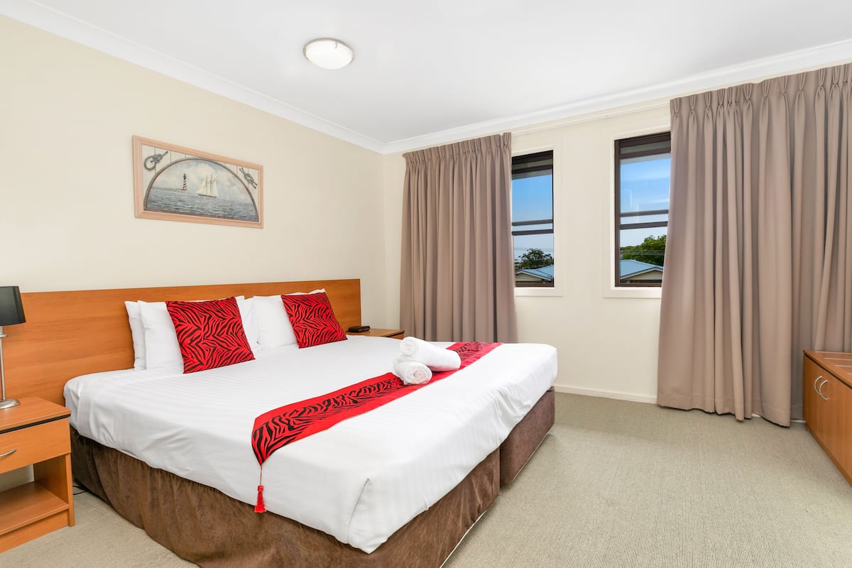 Een netjes opgemaakt bed met wit en rode beddengoed, een houten hoofdbord en een rode en zwarte gestreepte plaid. Er staat een nachtkastje met een lamp, een raam met beige gordijnen, en aan de muur hangt een schilderij van een zeilboot.