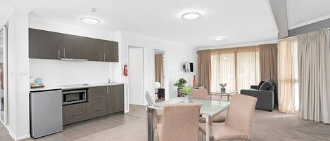 A modern kitchen with a dining area, a living room with a sofa, and a large window with curtains.