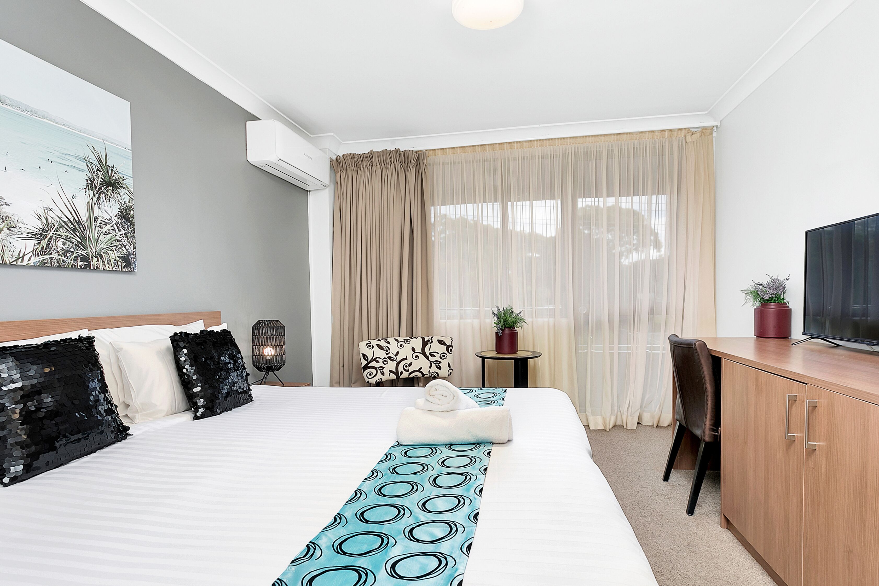 Quarto de hotel com uma cama grande, uma escrivaninha, uma cadeira, uma TV e uma janela com cortinas.