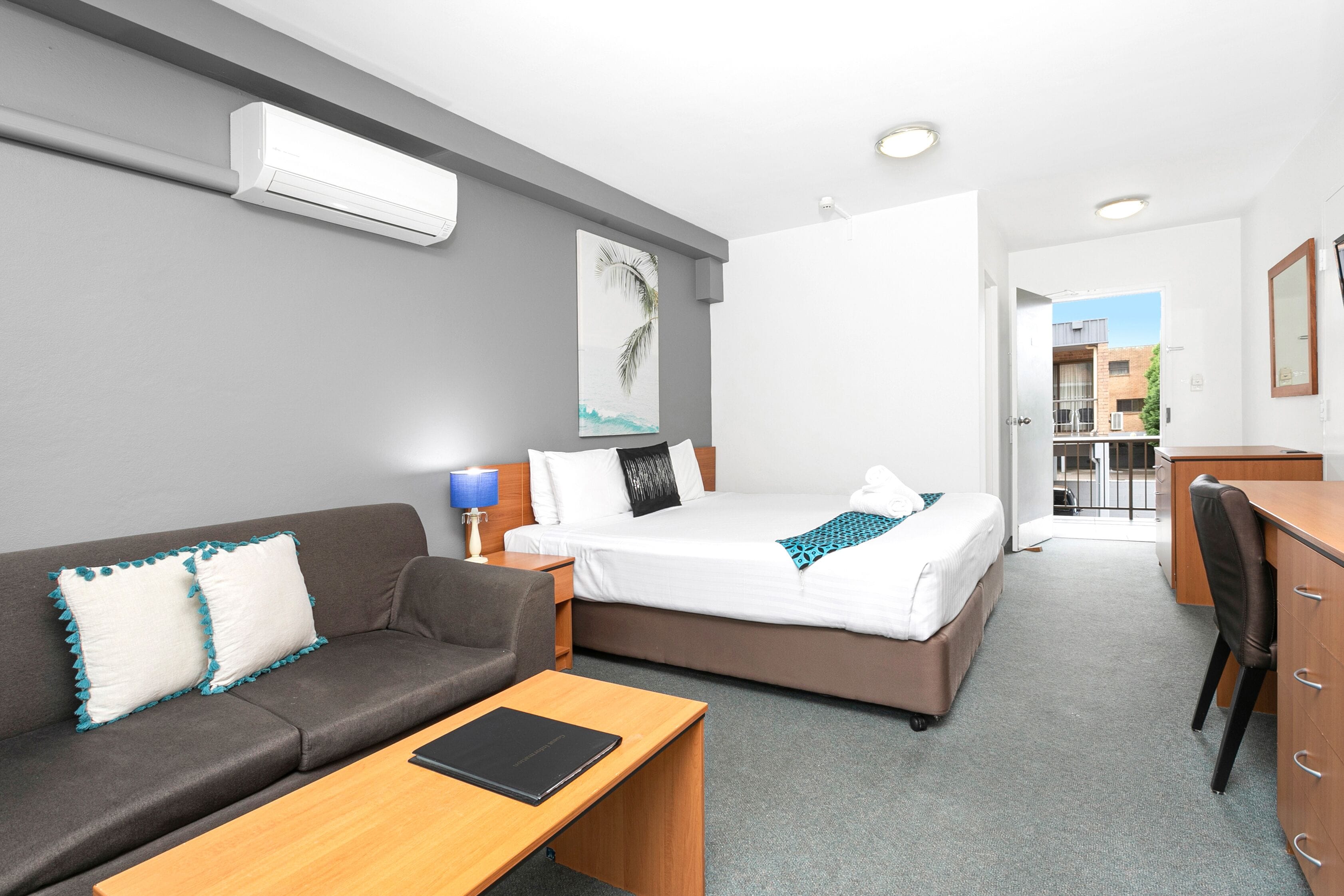 Et moderne hotelværelse med en grå sofa, et træskrivebord, en seng med hvide sengetæpper og en loftmonteret aircondition.