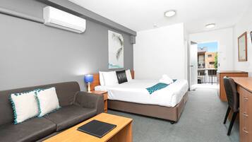 Et moderne hotelværelse med en grå sofa, et træskrivebord, en seng med hvide sengetæpper og en loftmonteret aircondition.