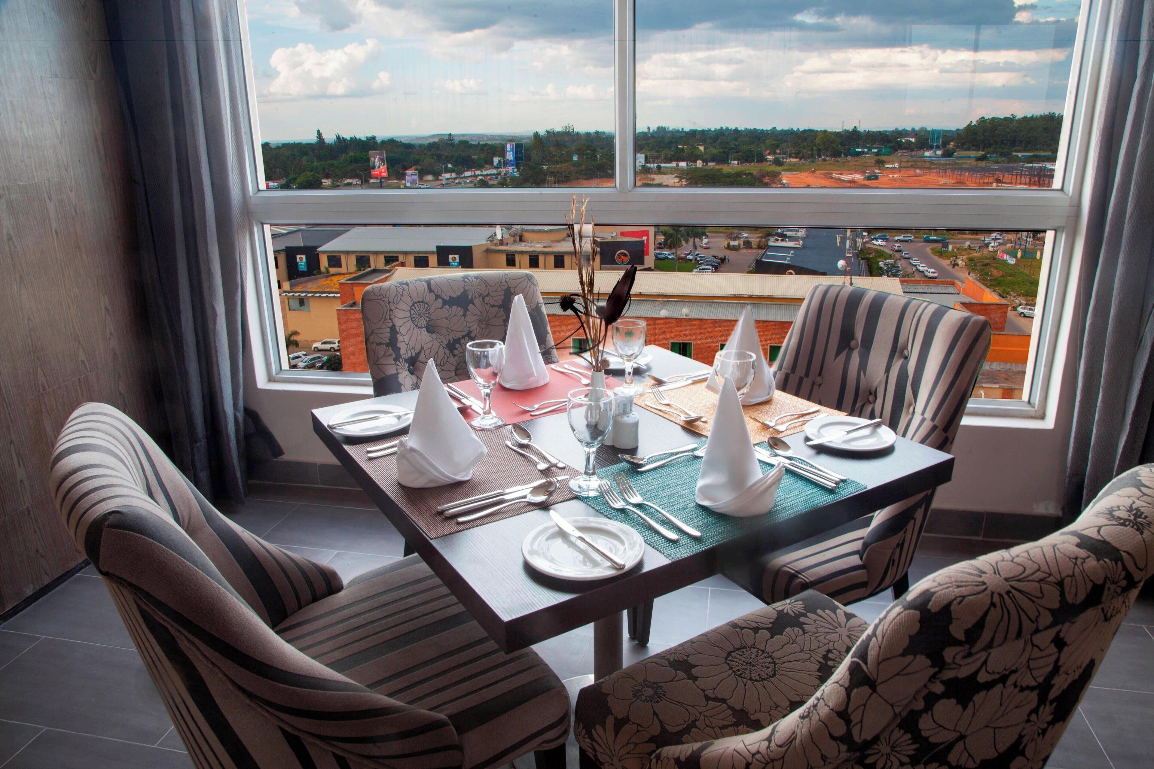 A dining area with a table set for four, chairs with striped and floral upholstery, and a view of a cityscape through large windows.