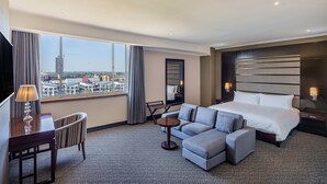 A hotel room with a large bed, a sofa, a desk, and a chair. There is a window with a view of buildings outside.