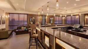 A modern hotel lobby with a central bar area, seating arrangements, and large windows with curtains.