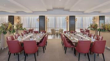 Un vaste espace dédié à la salle à manger, avec de longues tables et des chaises rouges, de grandes fenêtres ornées de rideaux et des plantes décoratives.