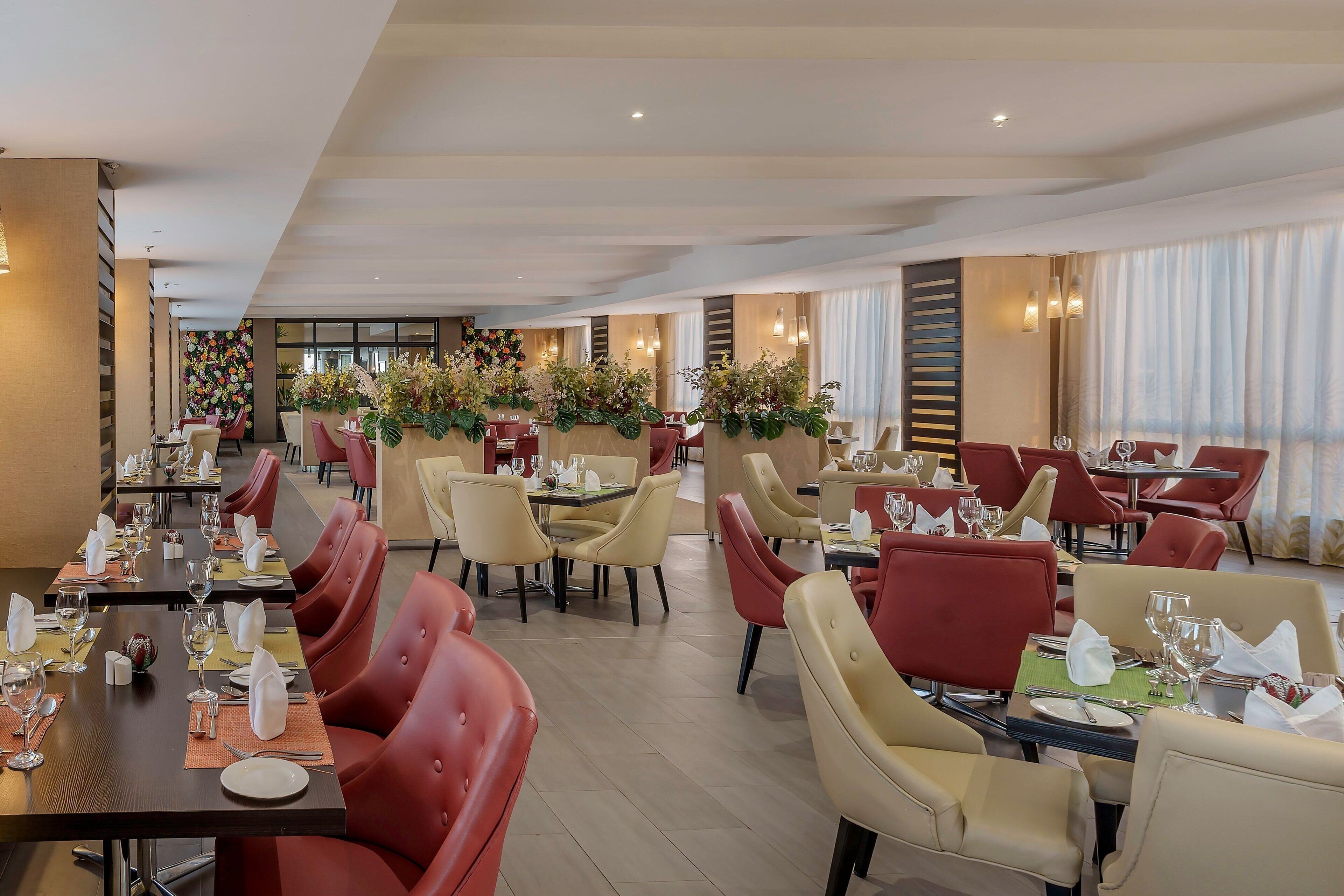 A modern dining area with red and beige chairs, neatly set tables, and decorative plants.