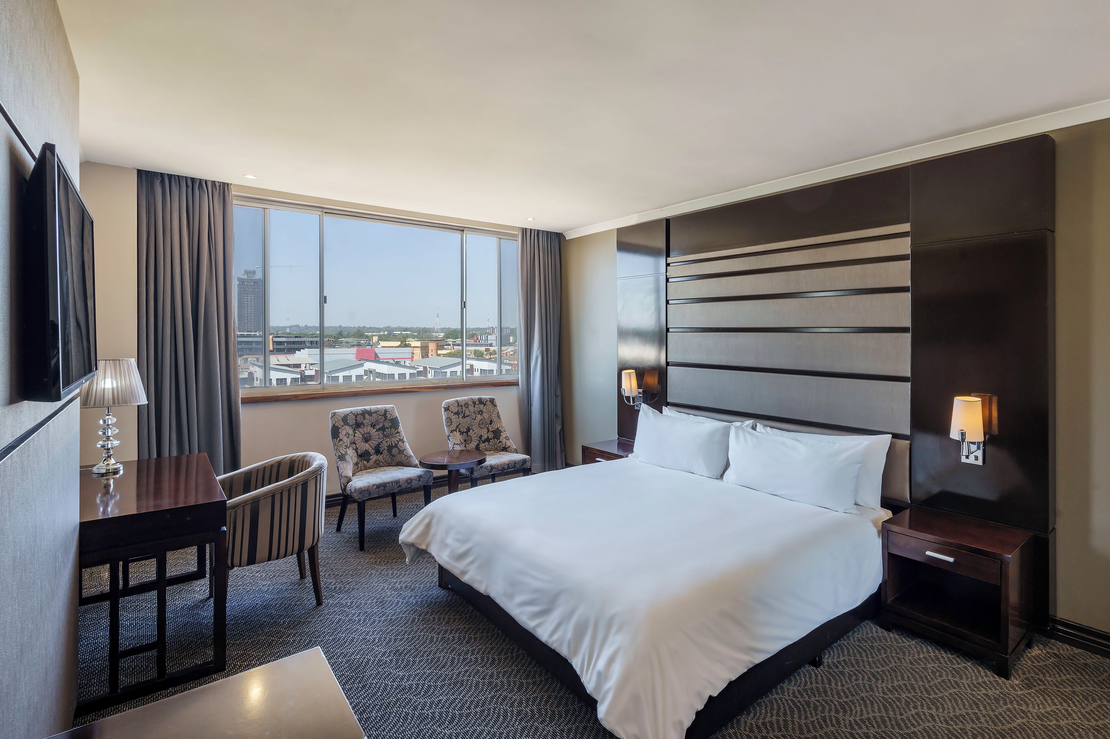 A hotel room with a large bed, a desk with a lamp, two chairs, a nightstand with a lamp, and a view of the city through large windows.