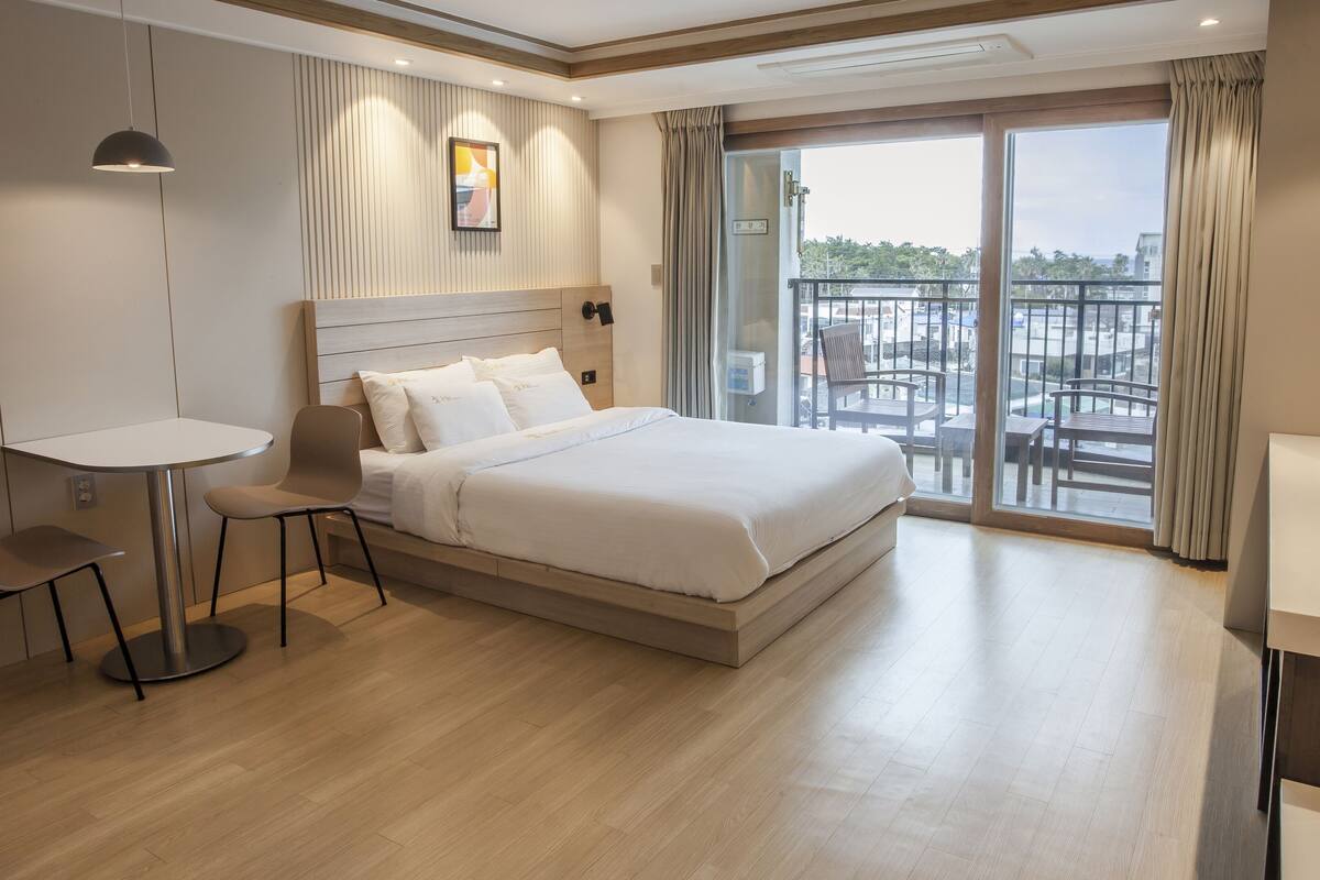 A modern hotel room with a large bed, a small table, a chair, and a balcony with a view.