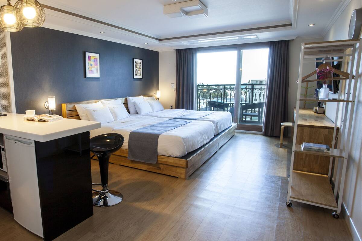 A modern hotel room with a large bed, a desk with a chair, a small kitchen area, and a balcony with a view.