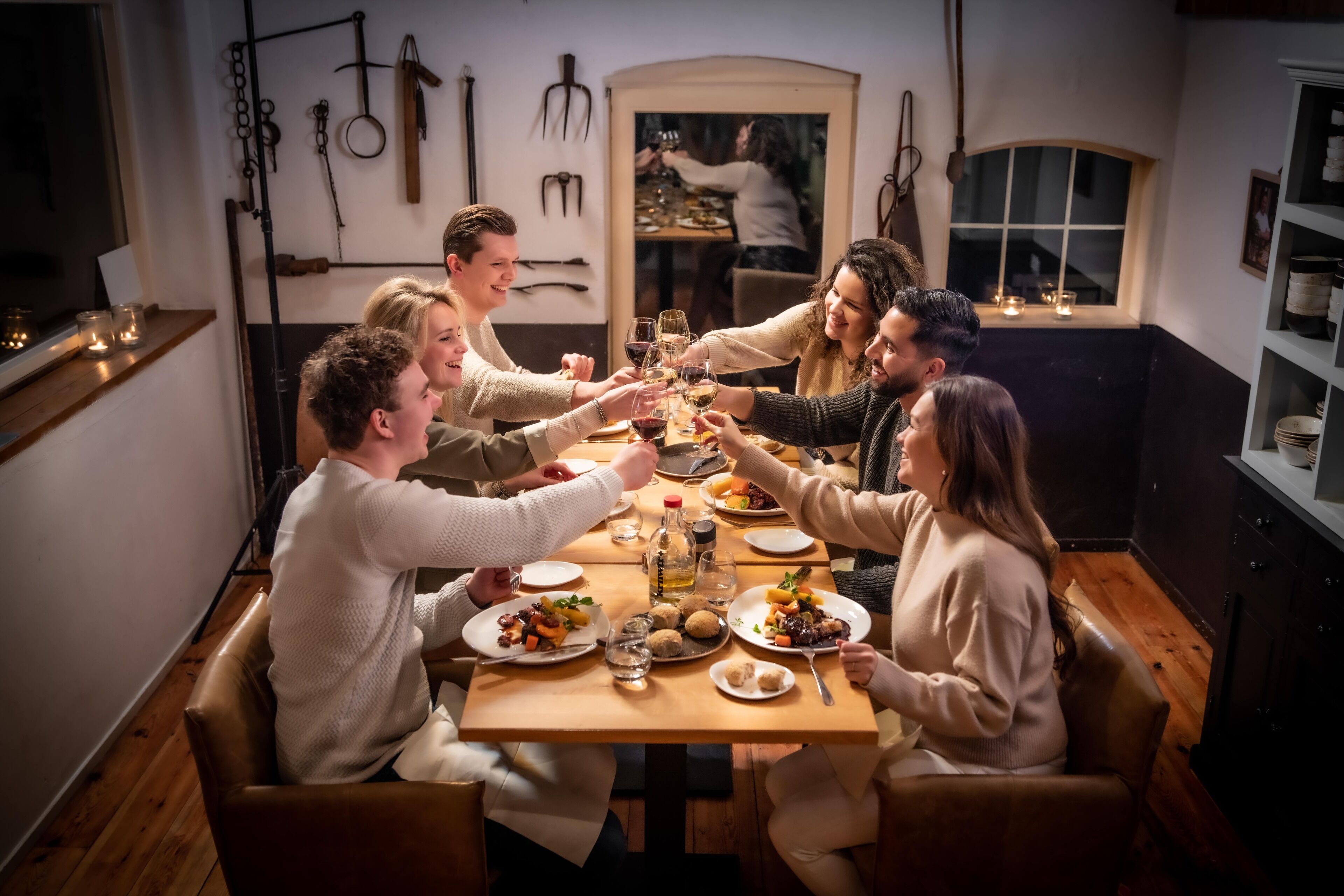 A group of people toasting at a wooden dining table in a warmly lit room with wooden floors and a window.