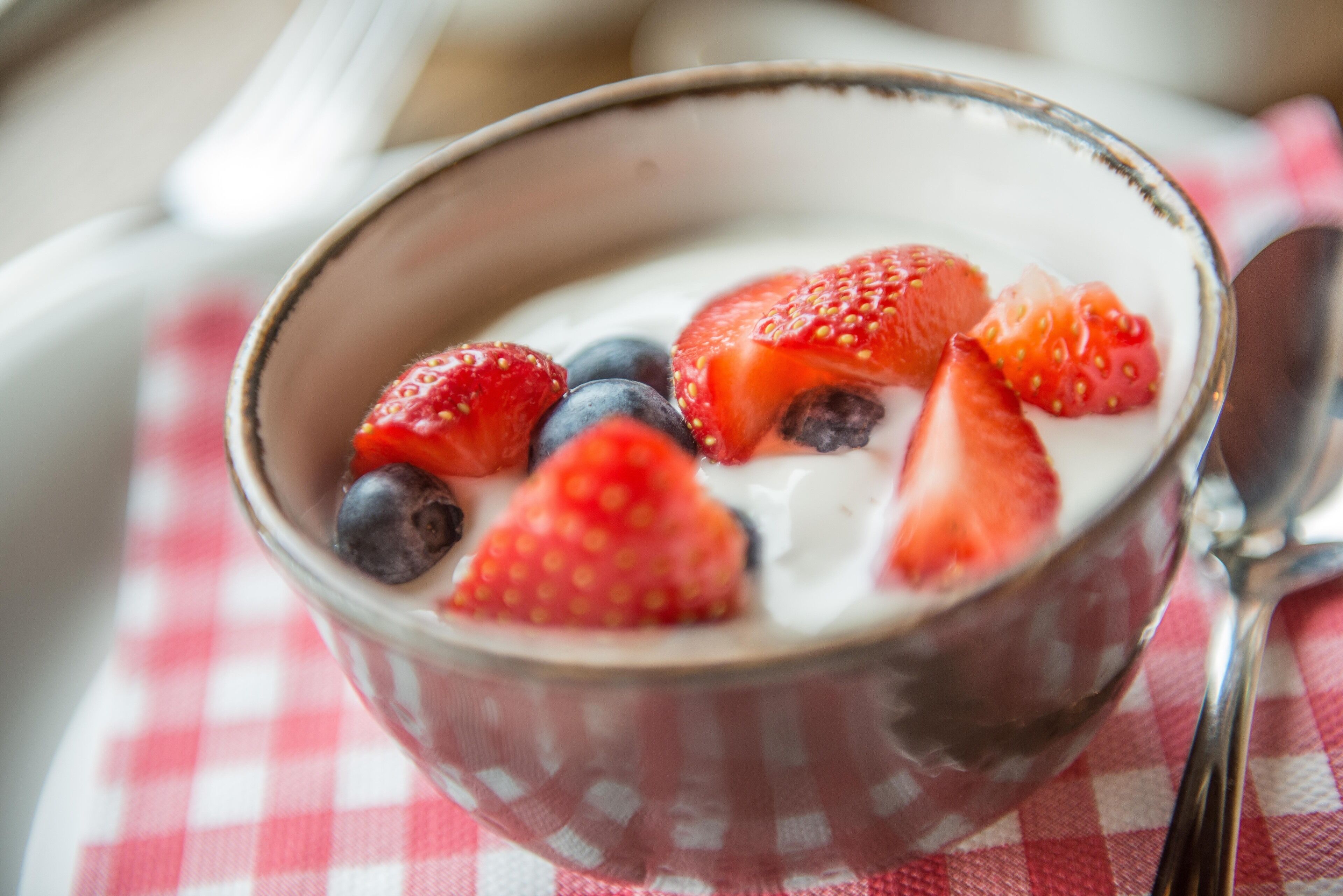 A bowl of yogurt with strawberries and blueberries on a checkered tablecloth.