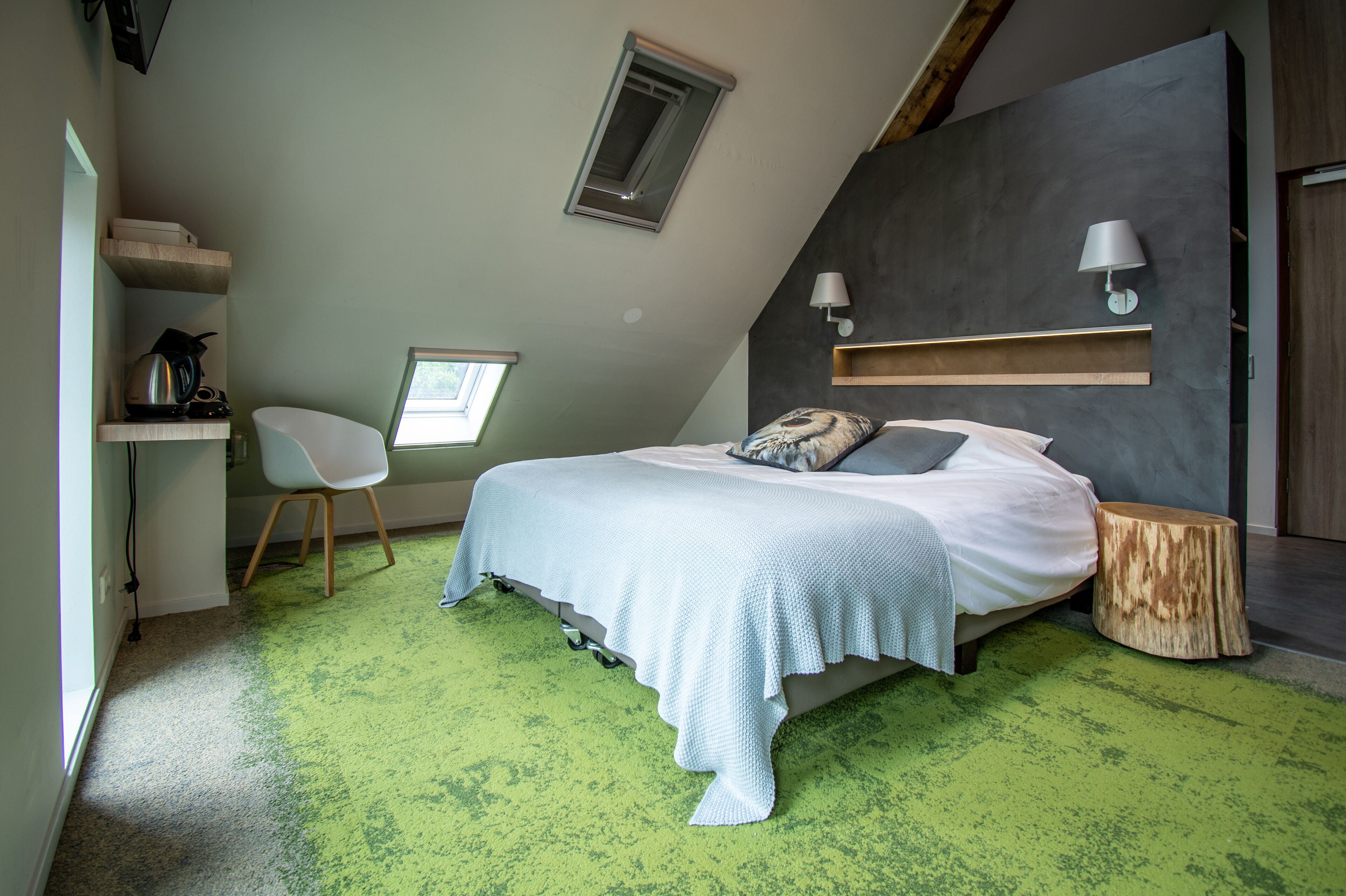 A bedroom with a sloped ceiling, a bed with a white bedspread, a white chair, a small table, and a wall-mounted lamp.